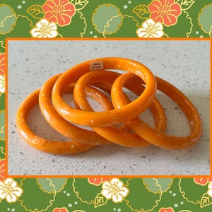 May include: A set of four smooth, glossy orange bangle bracelets with subtle white flecks throughout the material. One of the bracelets features a small white rectangular sticker with the black text "Made in Italy" printed on the inner surface.
