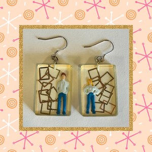 May include: A pair of earrings with clear resin and gold accents. Each earring features a miniature figure of a person standing in front of a stack of gold frames.