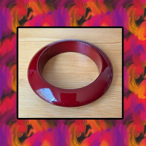 May include: A glossy, deep red bangle bracelet. The circular bracelet has a smooth, polished surface and a wide band. The jewelry is displayed on a light wood surface. The background is a colorful abstract design with shades of red, orange, and purple.
