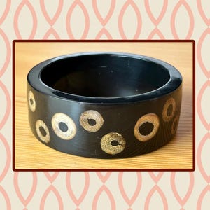 May include: A wide, circular black bangle bracelet featuring a series of inlaid, light-colored, textured circular patterns that resemble wood grain or bamboo cross-sections scattered across the outer surface of the dark, smooth material.