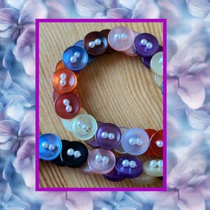 May include: A bracelet made of colorful buttons in shades of blue, brown, orange, purple, and white. Each button has two small, white pearl-like beads. The bracelet is set against a wooden background and framed in a purple border.