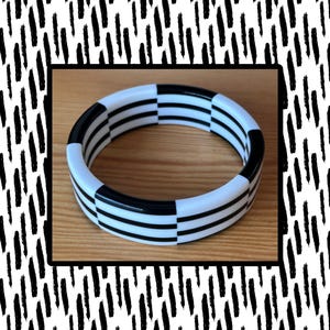 May include: A circular bangle bracelet featuring a repeating pattern of alternating black and white segments. The design consists of three horizontal black stripes running continuously around the circumference of the white and black bangle.