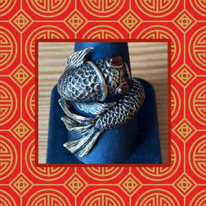 May include: A metallic ring shaped like a koi fish with a textured, dark silver-toned finish and two small, round, deep red stones set as eyes. The fish body wraps around the finger, featuring detailed scales and flowing fins on the tail end.
