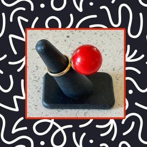 May include: A gold ring with a large, round, red bead. The ring is displayed on a black ring holder. The ring holder is rectangular with a rounded top. The background is a dark blue with white abstract shapes.
