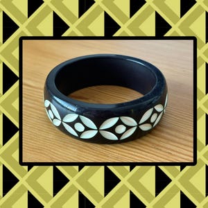 May include: A dark navy blue bangle bracelet featuring a repeating geometric pattern of inlaid white circular and petal-shaped elements. The smooth, rounded band has a polished finish and a consistent width, showcasing a clear contrast between the dark base and the light-colored decorative inlays.
