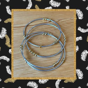 May include: Four silver-toned bangles with gold-colored beads are arranged on a light brown wooden surface. The bangles are inside a gold glitter frame, set against a black background with white and gold feather patterns. The jewelry is simple and elegant.