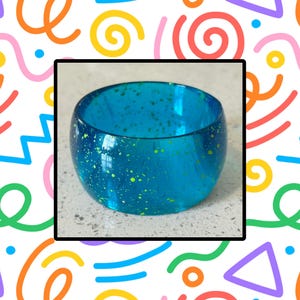 May include: A turquoise blue bangle bracelet with a speckled pattern of yellow-green dots. The bracelet is a solid, rounded shape, and appears to be made of a translucent material. The background is a colorful pattern of shapes and lines.