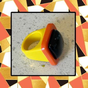 May include: A bold, geometric ring featuring a yellow band and an orange bezel. The ring holds a large, black, oval-shaped stone. The design is modern and eye-catching, perfect for adding a pop of color to any outfit.