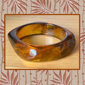 May include: A translucent amber-colored bangle bracelet with a smooth, polished finish and a rounded square shape. The material features swirling patterns of dark and light brown tones throughout, creating a marbled effect with light reflections.