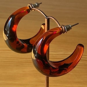 May include: A pair of hoop earrings with a tortoiseshell pattern. The earrings are a warm amber color with dark brown and black marbling. The earrings are displayed on a copper-colored earring stand.