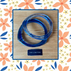 May include: Two blue resin bangles with a marbled effect, resting on a wooden surface. The bangles are a vibrant shade of blue and have a smooth, glossy finish. A small black tag with the text "THE LIMITED" is also present.