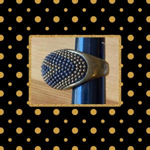 May include: A ring featuring a wide, rounded face covered in a dense pattern of small, dark blue and metallic gold-toned beads. The band of the ring is made of a smooth, antiqued brass-colored metal that contrasts with the textured, beaded surface.