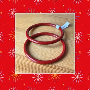 May include: Two red bangles are stacked on a wooden surface. The bangles are a solid, glossy red color. A small white tag is attached to one of the bangles. The background is a red pattern with white and yellow starburst designs.
