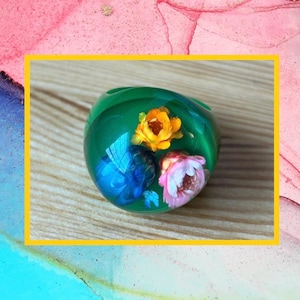 May include: A green resin ring with embedded flowers in shades of yellow, pink, and blue. The ring is set on a wooden surface, with a colorful abstract background.