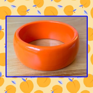 May include: A solid, bright orange bangle bracelet with a smooth, glossy finish and a thick, rounded profile. The bracelet features a simple, continuous circular design with no additional patterns, embellishments, or hardware visible on its surface.