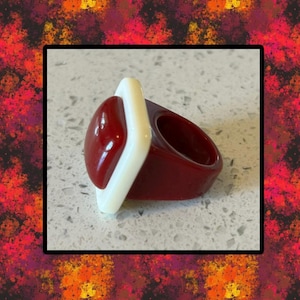 May include: A bold, statement ring featuring a deep red, oval-shaped center stone framed by a white rectangular accent. The ring's band is a matching deep red, creating a striking contrast. This unique piece is a fashion accessory.