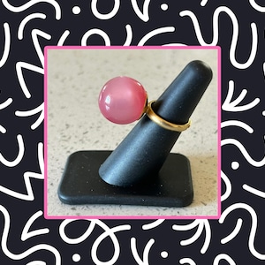 May include: A pink cabochon ring with a gold band. The ring features a large, round, pink stone. The ring is displayed on a black ring holder.