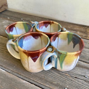 Puede incluir: Cuatro tazas de cerámica hechas a mano con patrones de esmalte coloridos únicos. Cada taza presenta un asa redondeada y un acento decorativo cerca de la base del asa. Las tazas muestran una mezcla de colores, incluyendo marrón, azul, amarillo y verde.
