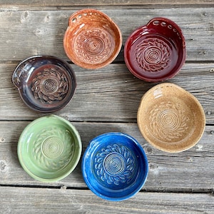 May include: Six handmade ceramic bowls in shades of brown, red, green, blue, and beige. Each bowl has a spiral and leaf pattern in the centre. The bowls are displayed on a weathered wooden surface.