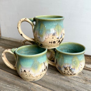 Puede incluir: Tres tazas de cerámica hechas a mano con un esmalte único. Las tazas tienen forma redondeada con asa y un esmalte moteado de dos tonos en tonos azules, verdes y beige. Las tazas están apiladas sobre una superficie de madera.