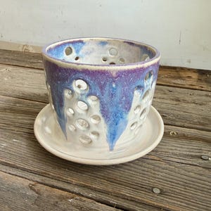 May include: A ceramic planter with a matching saucer, featuring a cylindrical shape and numerous circular cutouts. The exterior is finished with a glossy glaze in shades of white, blue, and purple, with the colours dripping down the sides.