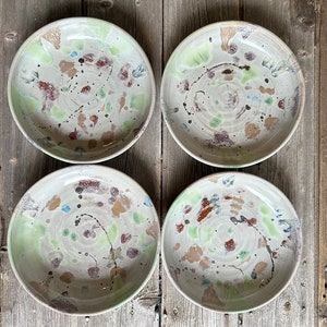 Könnte beinhalten: Vier handgefertigte Keramikschalen mit einem gesprenkelten Design. Die Schalen haben eine hellfarbige Basis mit grünen, braunen, blauen und schwarzen Spritzern. Die Schalen sind auf einer rustikalen Holzoberfläche angeordnet.