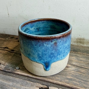 May include: A cylindrical ceramic planter featuring a two-tone glaze. The upper portion is finished in a vibrant blue with dark brown rim accents, while the lower section remains an unglazed, matte off-white clay with a visible drip pattern.