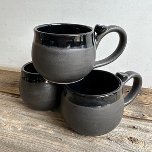 May include: Three black ceramic mugs with a rounded shape and a glazed finish. The mugs are stacked on top of each other.