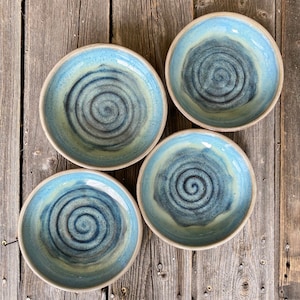 May include: Four handmade ceramic bowls with a blue and cream swirl design. The bowls have a light blue exterior and a darker blue spiral pattern in the center. The bowls are set on a rustic wooden surface.
