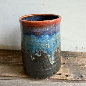 May include: A cylindrical ceramic vessel featuring a reactive glaze with a dark brown base that transitions into a vibrant blue midsection and a warm orange rim. The glaze exhibits a distinct drip pattern where the blue color overlaps the dark brown.