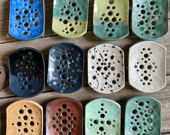 May include: Twelve rectangular ceramic soap dishes arranged in three rows. Each dish features a unique glaze color, including blue, green, black, white with black speckles, tan, and brown, and contains a pattern of circular drainage holes.