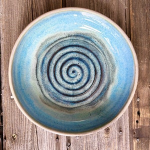 Könnte beinhalten: Eine handgefertigte Keramikschale mit hellblauer Glasur und einem Spiralmuster in der Mitte. Die Schale hat einen breiten Rand und eine strukturierte Oberfläche. Die Schale hat einen Durchmesser von etwa 25 cm, ideal zum Servieren von Salaten oder Pasta.