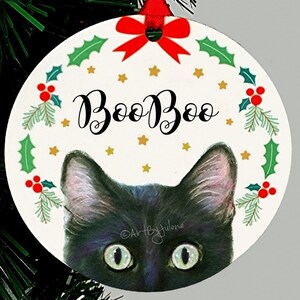 May include: A round Christmas ornament with a black cat peeking out from behind a wreath. The wreath is decorated with holly, berries, and gold stars. The ornament says "Boo Boo" in black cursive lettering.