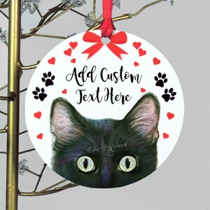 May include: A round white Christmas ornament with a black cat peeking out from the bottom. The ornament has a red bow, red hearts, and black paw prints around the edge. The text "Add Custom Text Here" is written in black on the ornament.