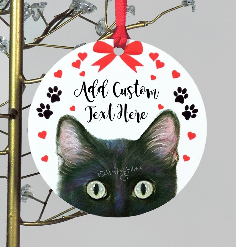May include: A round white Christmas ornament with a black cat peeking out from the bottom. The ornament has a red bow, red hearts, and black paw prints around the edge. The text "Add Custom Text Here" is written in black on the ornament.