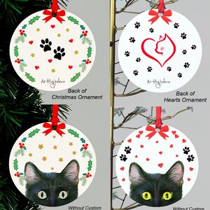 May include: Four round Christmas ornaments with a black cat peeking out from behind a wreath of holly and stars. The ornaments are white with red hearts and paw prints. The back of the ornaments show different designs, including a wreath of holly and stars, a heart with a cat inside, and a cat's face.