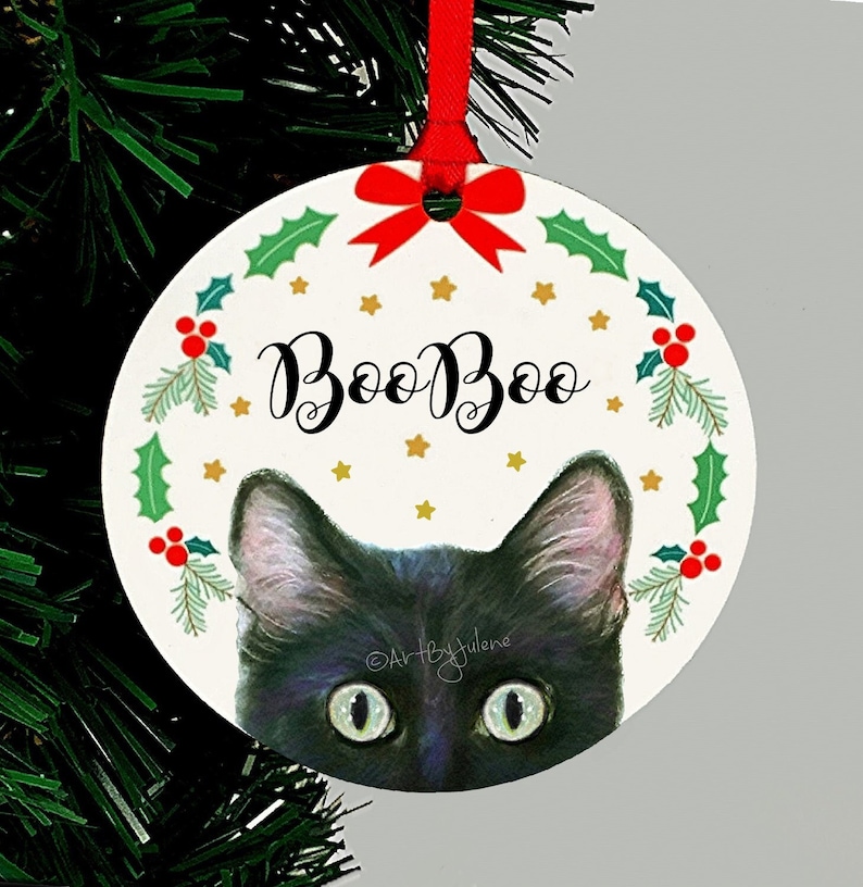 May include: A round Christmas ornament with a black cat peeking out from behind a wreath. The wreath is decorated with holly, berries, and gold stars. The ornament says "Boo Boo" in black cursive lettering.