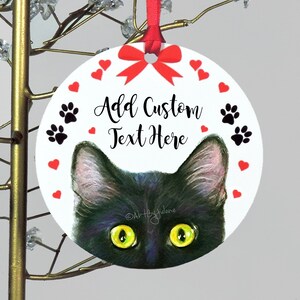 May include: Round white Christmas ornament with a black cat peeking out from the bottom. The ornament has a red bow, red hearts, and black paw prints around the edge. The text "Add Custom Text Here" is written in black cursive.