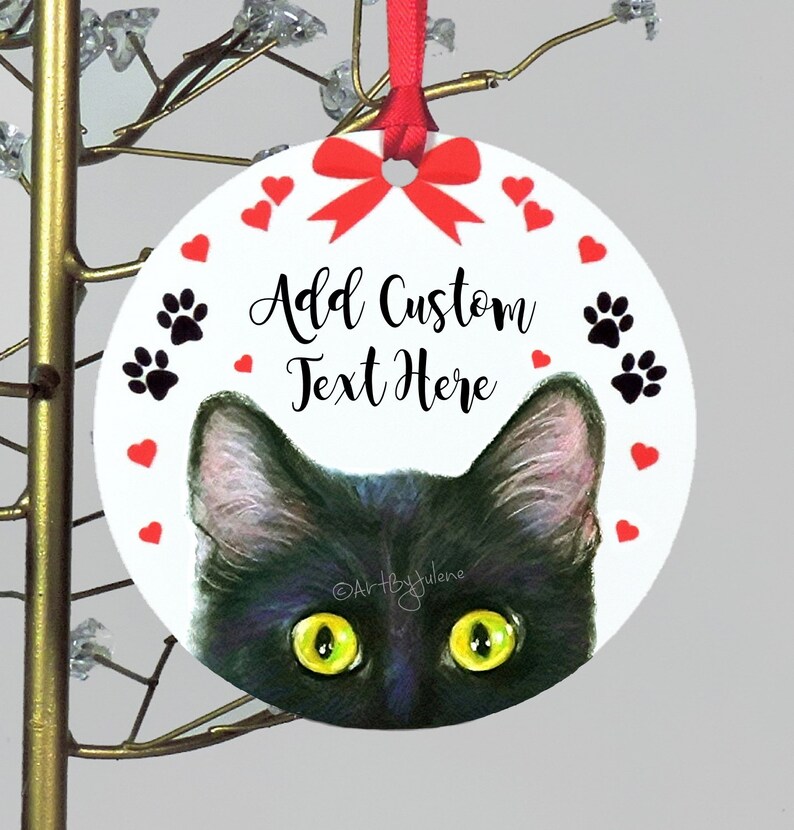 May include: Round white Christmas ornament with a black cat peeking out from the bottom. The ornament has a red bow, red hearts, and black paw prints around the edge. The text "Add Custom Text Here" is written in black cursive.