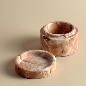 May include: A cylindrical stone container with a matching circular lid placed beside it. The object is crafted from polished pink and peach marble, featuring natural dark brown veining and a smooth, glossy finish throughout its surface.