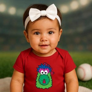 May include: A baby wearing a red onesie with a colorful monster graphic and a white bow headband. A baseball is in the background. The baby is sitting on a cream-colored blanket.