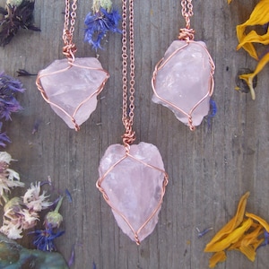 May include: Three rose quartz crystal pendants wrapped in copper wire, hanging from delicate copper chains. The crystals are a pale pink color and have a natural, rough texture. The pendants are displayed against a wooden background.