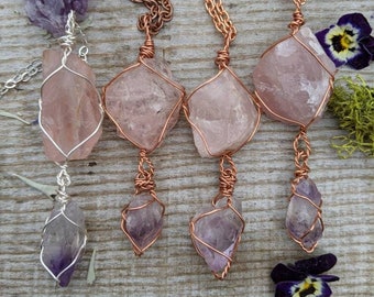 Puede incluir: Tres collares envueltos en alambre de cobre con colgantes de cristal de diferentes colores. Los colliers presentan un colgante de cuarzo rosa, una amatista y un colgante de cuarzo rosa más grande.