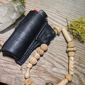 Puede incluir: Una funda de cuero negro para encendedor con un encendedor plateado en su interior. La funda est&aacute; unida a un collar de cuentas de madera natural con intrincados tallados.