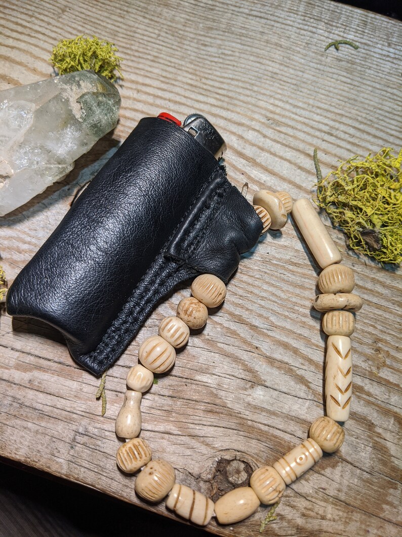 Puede incluir: Una funda de cuero negro para encendedor con un encendedor plateado en su interior. La funda est&aacute; unida a un collar de cuentas de madera natural con intrincados tallados.