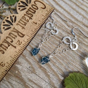 May include: Silver earrings with a chain and a blue crystal pendant. The earrings are hanging from a wooden ruler with the words "Craftsman Rule" printed on it.