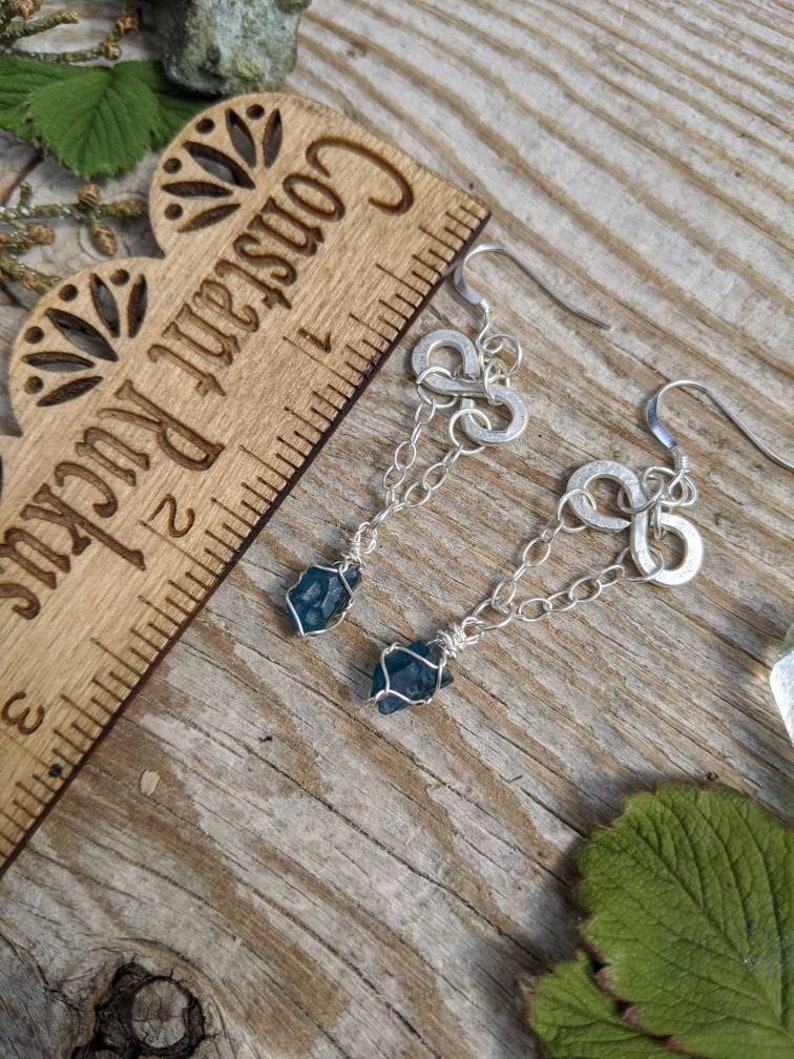 May include: Silver earrings with a chain and a blue crystal pendant. The earrings are hanging from a wooden ruler with the words "Craftsman Rule" printed on it.