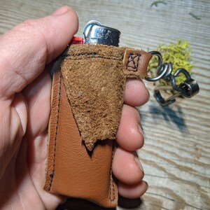 May include: A brown leather keychain with a small pocket for a lighter. The lighter is silver with a red plastic top. The keychain has a metal loop and a chain.