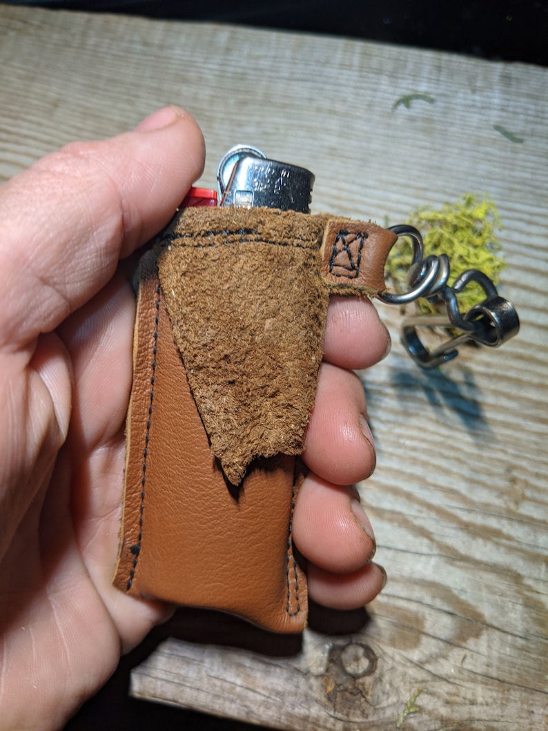 May include: A brown leather keychain with a small pocket for a lighter. The lighter is silver with a red plastic top. The keychain has a metal loop and a chain.