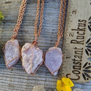 May include: Three pink crystal pendants wrapped in copper wire, hanging from copper chains. The pendants are approximately 1 inch long and are displayed on a wooden surface with a ruler.